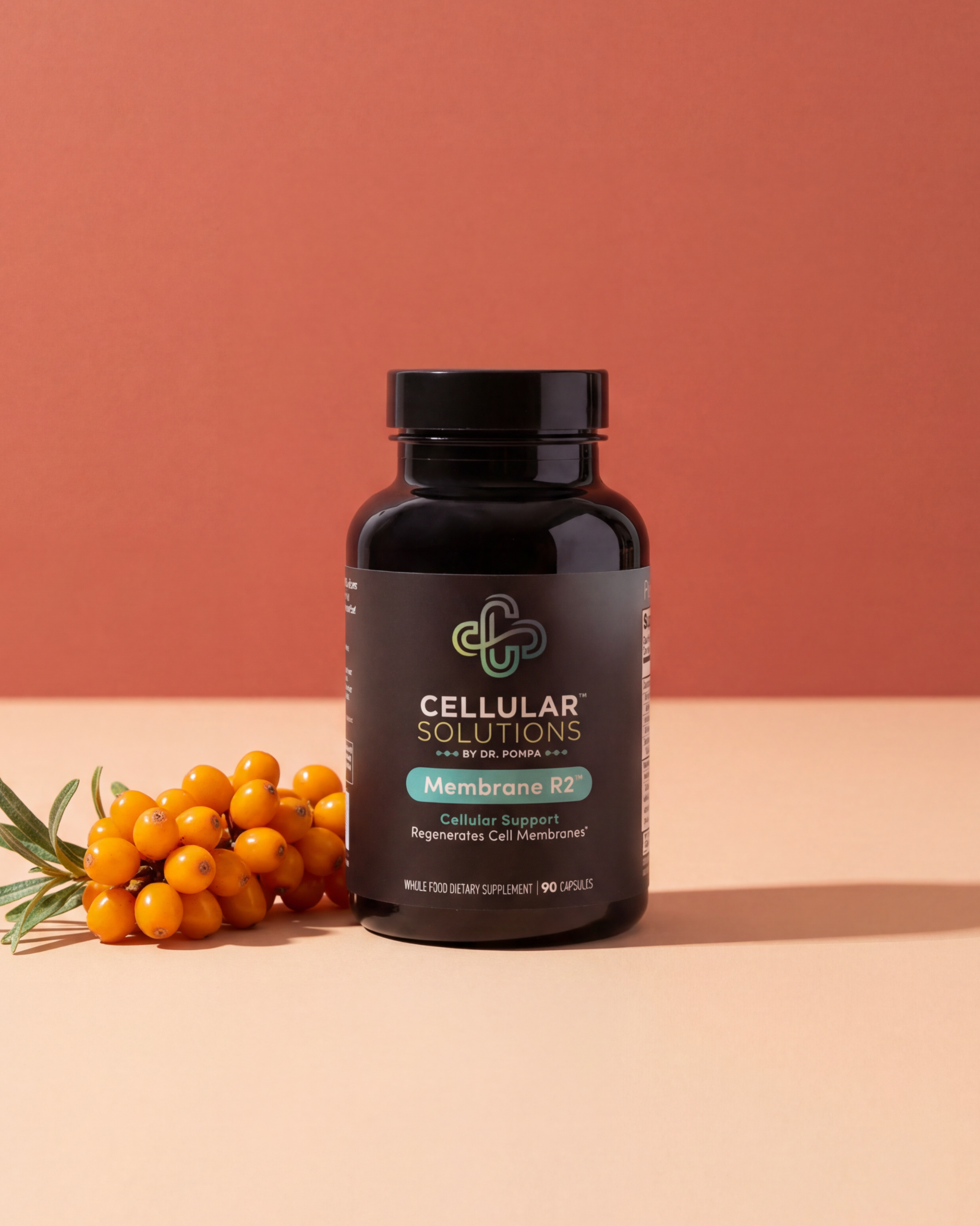 Cellular Solutions Membrane R2 bottle on a deep clay backdrop with a sprig of fresh sea buckthorn berries beside it.